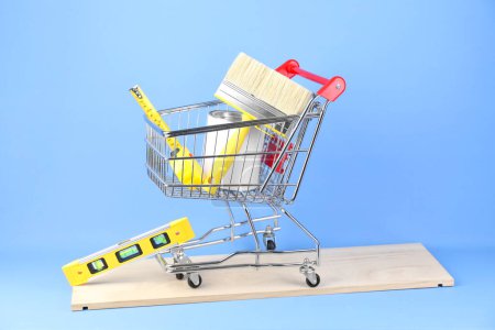 Foto de Pequeño carrito de compras con pintura y equipo de renovación sobre fondo azul claro - Imagen libre de derechos