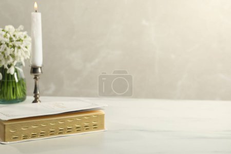 Burning church candle, Bible and flowers on white marble table. Space for text