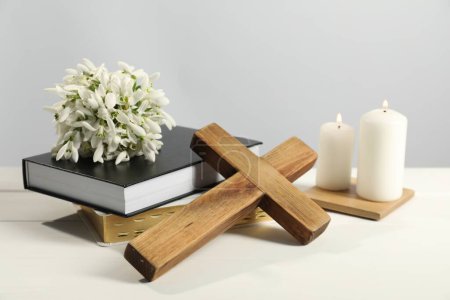 Burning church candles, wooden cross, ecclesiastical books and flowers on white table