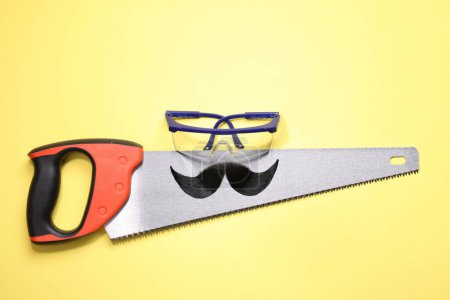 Téléchargez les photos : Visage d'homme fait de moustache artificielle, lunettes de sécurité et scie à main sur fond jaune, vue de dessus - en image libre de droit