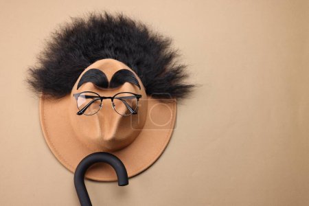 Téléchargez les photos : Visage d'homme en cheveux artificiels, sourcils, lunettes et chapeau sur fond beige, vue de dessus. Espace pour le texte - en image libre de droit