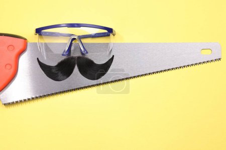 Téléchargez les photos : Visage d'homme fait de moustache artificielle, lunettes de sécurité et scie à main sur fond jaune, vue de dessus. Espace pour le texte - en image libre de droit