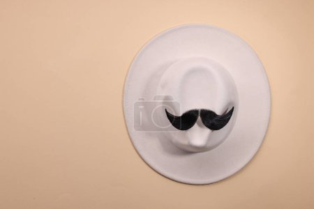 Téléchargez les photos : Visage d'homme fait de moustache artificielle et chapeau sur fond beige, vue de dessus. Espace pour le texte - en image libre de droit