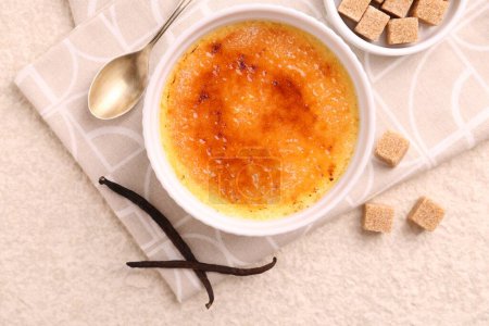 Foto de Deliciosa crema brulee en tazón, vainas de vainilla, cubos de azúcar y cuchara en la mesa de textura ligera, vista superior - Imagen libre de derechos