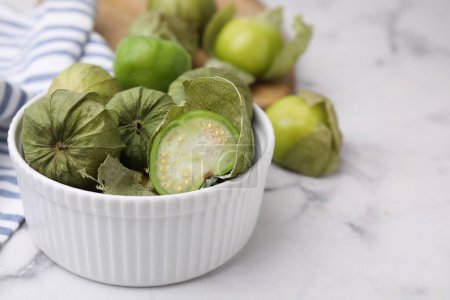 Fresh green tomatillos with husk in bowl on light marble table, closeup