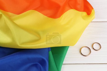 Rainbow LGBT flag and wedding rings on white wooden table, top view
