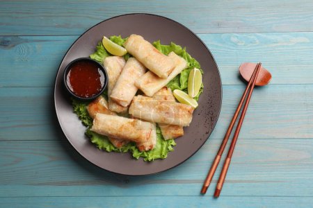 Plate with tasty fried spring rolls, lettuce, lime, sauce and chopsticks on light blue wooden table, top view