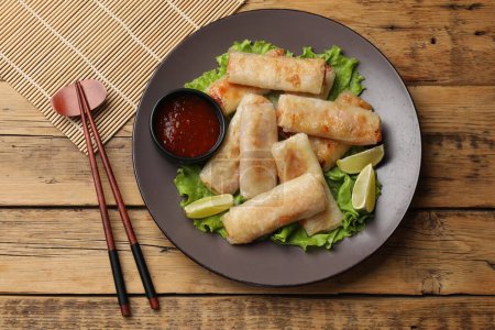 Plate with tasty fried spring rolls, lettuce, lime, sauce and chopsticks on wooden table, top view