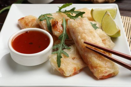 Tasty fried spring rolls, arugula, lime and sauce on table, closeup