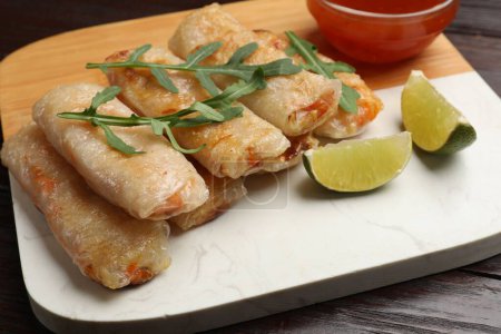 Tasty fried spring rolls, lime, arugula and sauce on wooden table, closeup