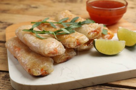 Tasty fried spring rolls, lime, arugula and sauce on wooden table, closeup