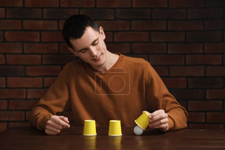 Shell game. Man showing ball under cup at wooden table