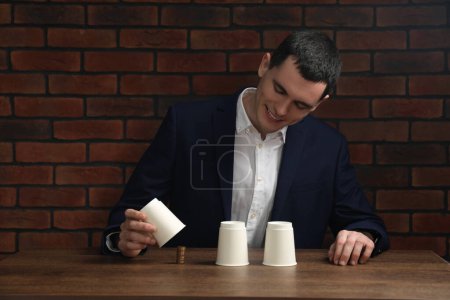 Shell game. Man showing stack of coins under cup at wooden table
