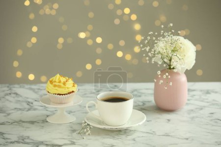 Delicious cupcake with yellow cream, coffee and flowers on white marble table against blurred lights