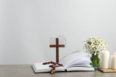 Kirchenkerzen, Bibel, Holzkreuz, Rosenkranz und Blumen auf grauem Tisch. Raum für Text