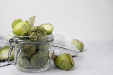 Fresh green tomatillos with husk in glass jar on light table, space for text