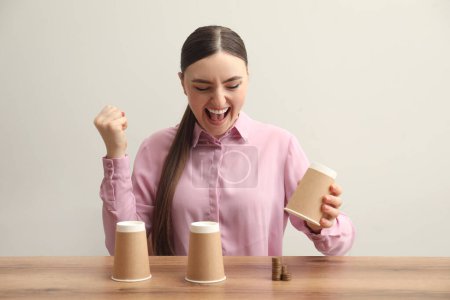 Shell game. Emotional woman showing stack of coins under cup at wooden table