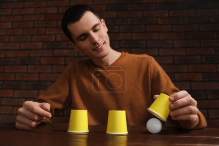 Shell game. Man showing ball under cup at wooden table, low angle view