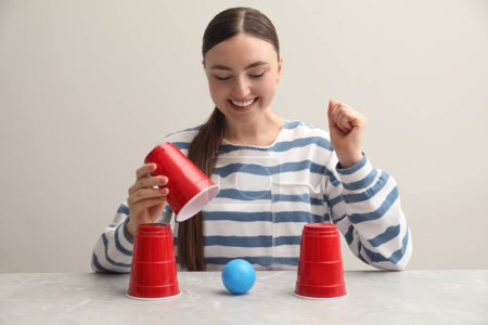 Shell game. Happy woman showing ball under cup at light marble table