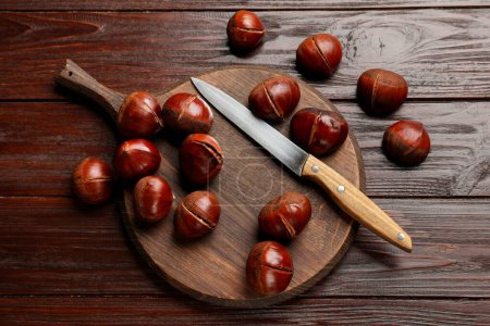 Roasted edible sweet chestnuts and knife on wooden table, flat lay