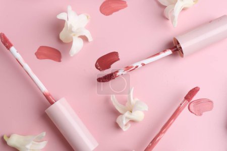 Foto de Diferentes brillos labiales, aplicadores y flores sobre fondo rosa, disposición plana - Imagen libre de derechos