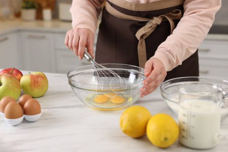 Woman whisking eggs in bowl at light marble table indoors, closeup