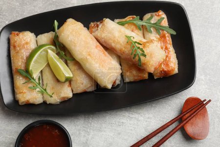Tasty fried spring rolls, arugula, lime and sauce served on grey textured table, flat lay
