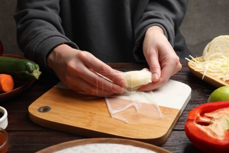 Making delicious spring rolls. Woman wrapping fresh cabbage into rice paper at wooden table, closeup