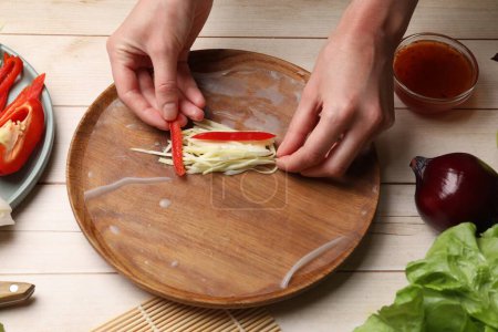 Making delicious spring rolls. Woman wrapping fresh vegetables into rice paper at wooden table, closeup