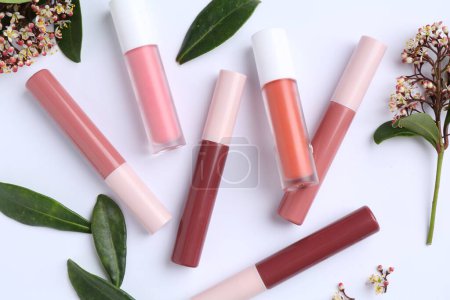 Different lip glosses, green leaves and flowers on white background, flat lay
