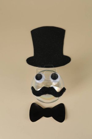 Téléchargez les photos : Visage d'homme en fausse moustache, boule, chapeau, noeud papillon et yeux sur fond beige, vue de dessus - en image libre de droit