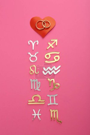 Foto de Compatibilidad con el zodiaco. Signos, anillos de boda y corazón rojo sobre fondo rosa, planas - Imagen libre de derechos