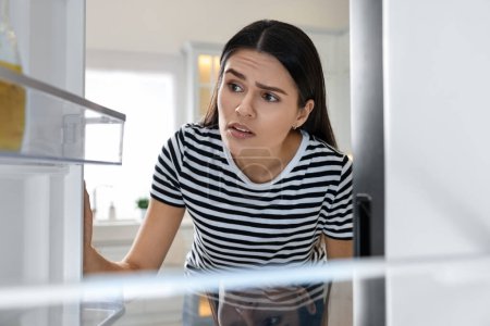 Mujer molesta cerca de refrigerador vacío en la cocina, vista desde el interior