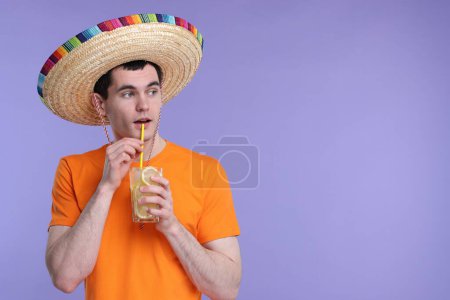 Joven con sombrero mexicano bebiendo cóctel sobre fondo violeta. Espacio para texto