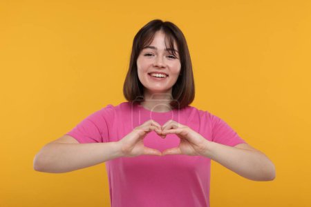 Téléchargez les photos : Femme heureuse montrant geste de coeur avec les mains sur fond orange - en image libre de droit