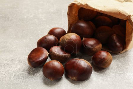 Paper bag with roasted edible sweet chestnuts on light grey table, closeup