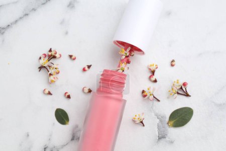 Rosafarbener Lipgloss, Blüten und grüne Blätter auf weißem Marmortisch, flache Lage