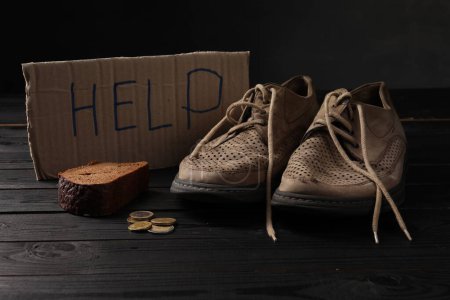 Téléchargez les photos : La pauvreté. Panneau en carton avec mot Aide, vieilles chaussures, morceau de pain et pièces de monnaie sur table en bois noir - en image libre de droit