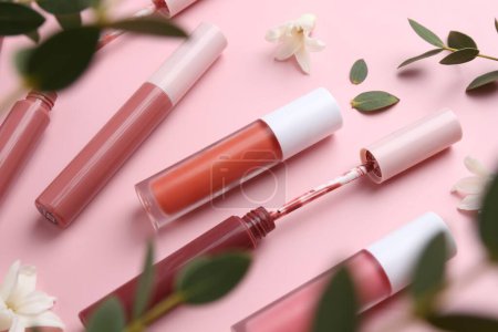 Foto de Brillos labiales diferentes, aplicador, flores y hojas verdes sobre fondo rosa - Imagen libre de derechos