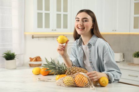 Téléchargez les photos : Femme avec citron et sac à ficelle de fruits frais à table en marbre clair dans la cuisine - en image libre de droit