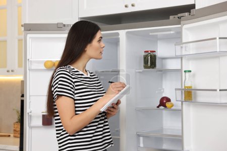 Mujer joven escribiendo notas cerca de refrigerador vacío en la cocina