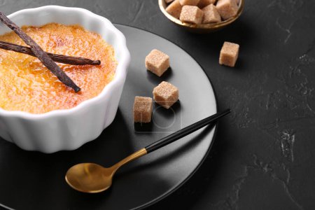 Foto de Deliciosa crema brulee en tazón, vainas de vainilla, terrones de azúcar y cuchara en la mesa de textura gris oscuro, primer plano - Imagen libre de derechos