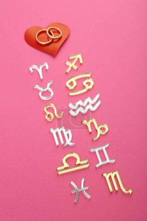 Foto de Compatibilidad con el zodiaco. Signos, anillos de boda y corazón rojo sobre fondo rosa, vista superior - Imagen libre de derechos