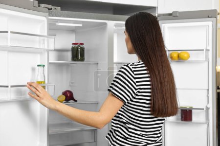 Mujer joven cerca de refrigerador vacío en la cocina, vista trasera