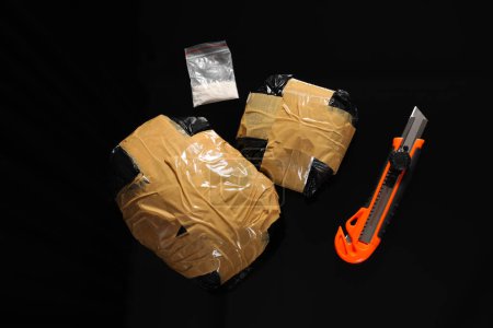 Smuggling, drug trafficking. Packages with narcotics and utility knife on black surface, flat lay