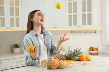 Téléchargez les photos : Sac à ficelle avec fruits frais sur table en marbre clair. Femme jetant du citron dans la cuisine - en image libre de droit
