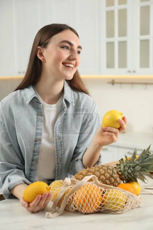 Téléchargez les photos : Femme avec citrons et sac à ficelle de fruits frais à table en marbre clair dans la cuisine - en image libre de droit