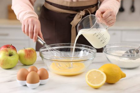 Woman whisking eggs while pouring milk into bowl at light marble table indoors, closeup