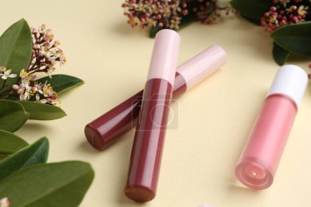 Different lip glosses, green leaves and flowers on pale yellow background, closeup