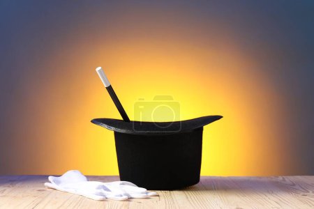 Magician's hat, wand and gloves on wooden table against color background